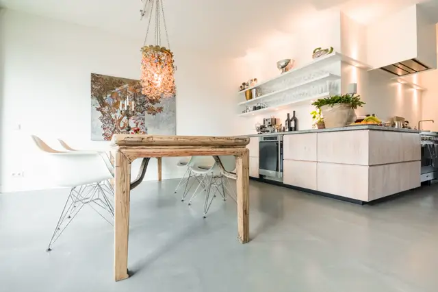 Ruime, modern ingerichte keuken met strakke witte wanden, houten tafel en naadloze vloer in licht grijs. Het licht valt zacht naar binnen en creëert een serene, opgeruimde sfeer.
