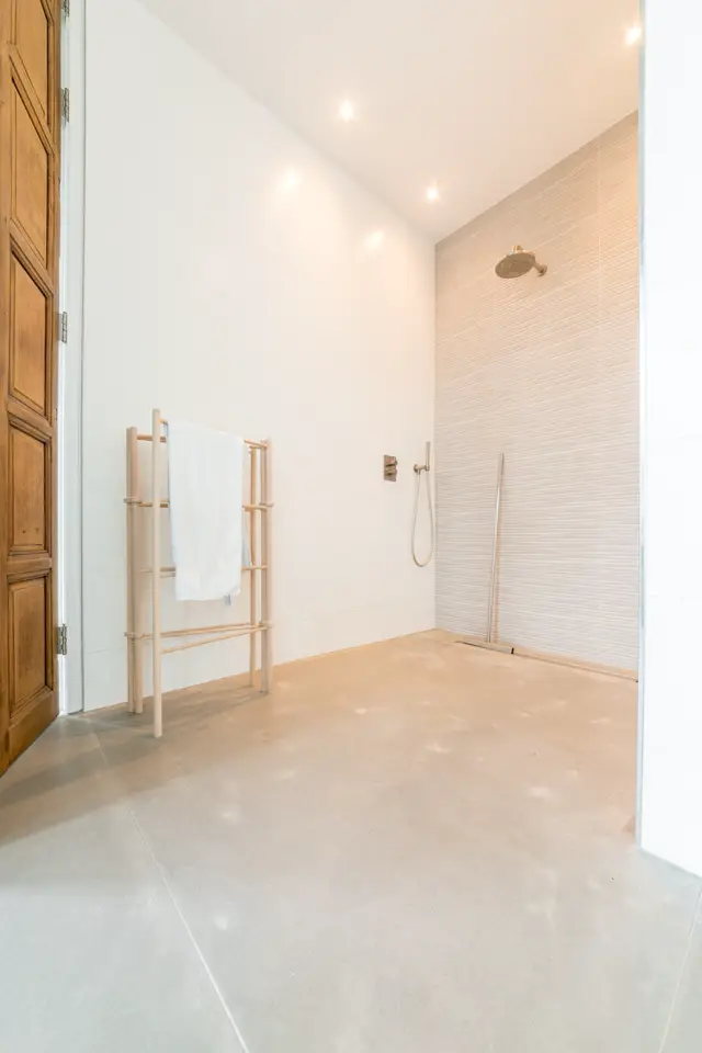 Ruime, lichte badkamer met naadloze vloer en wand. Warm gebroken wit en beige tonen, schoon design met houtaccenten en geborstelde armaturen.