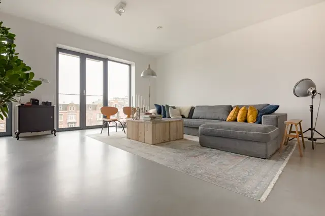 Ruim, licht woonkamer met grote raampartijen, grijze naadloze vloer en minimalistische inrichting. Scandinavische meubelen, gele accenten en strakke architectuur creëren een serene luxe sfeer.