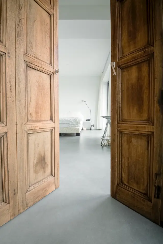 Een elegante doorgang met authentieke houten deuren aan beide zijden, uitkijkend op een serene slaapkamer met naadloze lichte vloer. Het contrast tussen warme houtaccenten en strakke witte wanden creëert een verfijnd ruimtelijk moment.