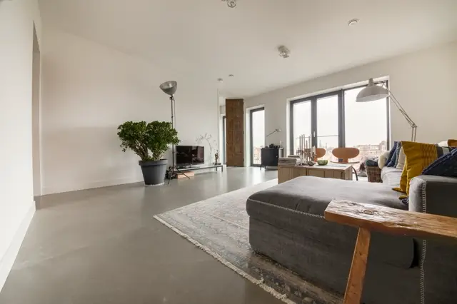 Ruime, lichte woonkamer met strakke naadloze vloer in warm grijs. Groot raamfront met stadsuitzicht, minimalistisch ingericht met loungezetel, houten accenten en perfect gebalanceerde styling.