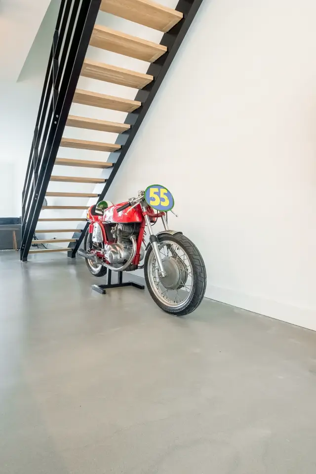 Moderne trap met houten treden en zwart metalen reling tegen witte wand. Daaronder een glimmende naadloze vloer met minimalistisch interieur en een rode vintage motorfiets als statement piece.