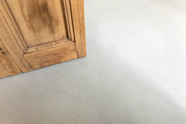 Detail van een naadloze lichte vloer die elegant aansluit op een klassieke houten deur. De minimalistische overgang benadrukt de zuiverheid van het ontwerp en de vakmanschap.