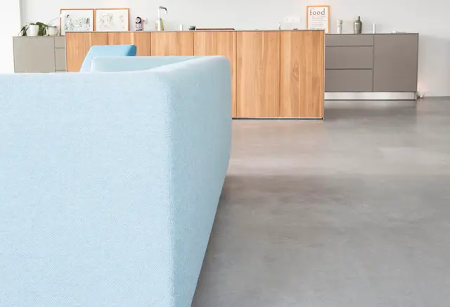 Minimalistisch ingerichte woonkamer met een lichtblauwe zithoek op een grijze naadloze vloer. Op de achtergrond houten kasten en neutrale wandelementen creëren een rustige, geordende ruimte.