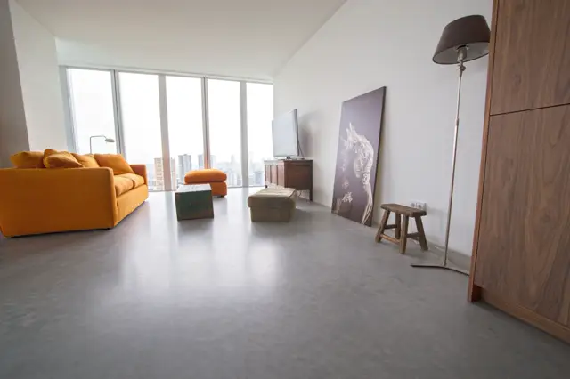 Ruime, lichte woonkamer op grote hoogte met een strakke naadloze vloer in licht grijs. Minimalistisch ingericht met een oranje sofa, houten accenten en panoramisch uitzicht over de stad.