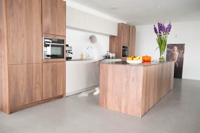 Een strakke, lichte keukenruimte met houten kasten, witte wanden en een naadloze grijze vloer. Het eiland domineert met warme houtnerf, aangevuld met verse bloemen en minimalistisch aanrecht.