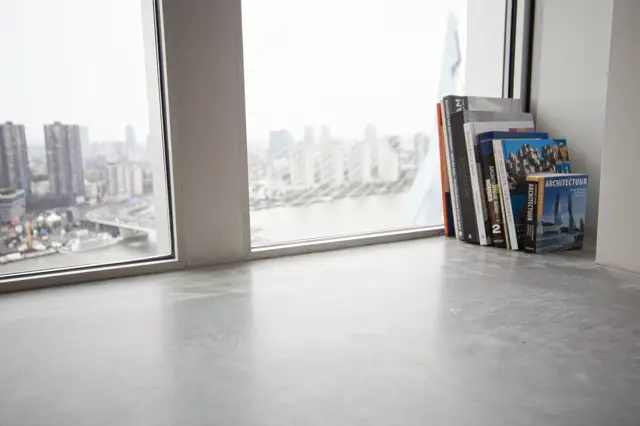 Een strakke naadloze vloer in lichte grijstint vormt de basis van deze high-rise woonruimte. Grote raampartijen bieden een panoramisch uitzicht op de skyline, terwijl enkele architectuurboeken subtiel de ruimte afmaken.