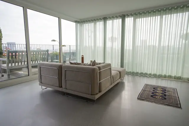 Lichte, ruime woonkamer met grote raampartijen en groene vitrage. Een strakke grijze naadloze vloer vormt het perfecte canvas voor de sobere inrichting met moderne meubels en uitzicht op het water.