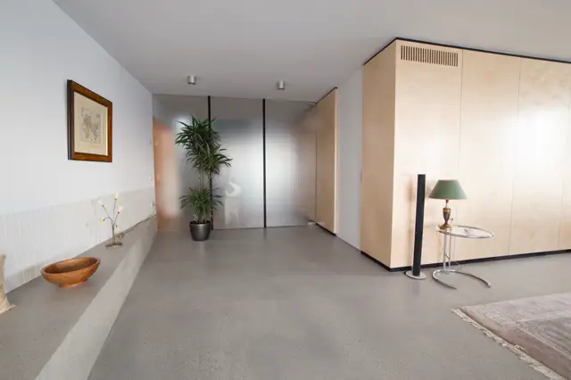 Serene slaapkamer met een grijze naadloze vloer die naadloos aansluit op een lichte, opgeruimde ruimte. Natuurlijke belichting en strakke architectuur met houten accenten creëren een rustgevend, luxe interieur.
