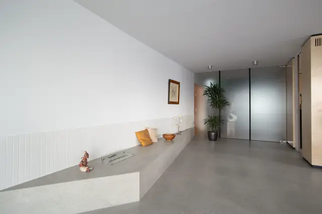 Een serene, vrijwel lege hal met een grijze naadloze vloer en witte paneelwand. Subtiele styling met gouden accenten, een plant en een strak glazen schuifdeur.