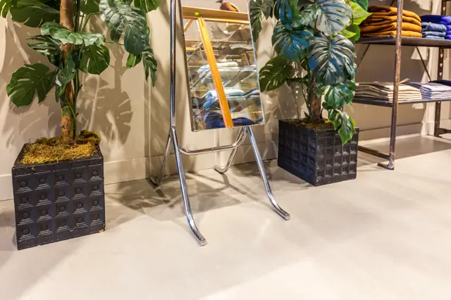 Een serene showroom met een lichte, naadloze vloer waarop zwarte plantenbakken met tropische planten zijn geplaatst. Natuurlijk zonlicht creëert scherpe schaduwen, ondersteund door minimalistisch interieur met opberging op achtergrond.