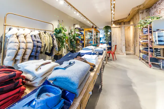Schone, moderne winkelruimte met lichte naadloze vloer en strak verlicht plafond. De ruimte toont kledingdisplays met houten elementen en groene planten in een opgeruimde, luxe setting.