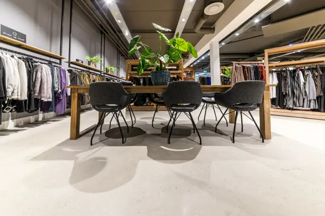 Ruime winkelruimte met lichte, naadloze vloer waarin subtiele patroondetails zijn geïntegreerd. Strakke, moderne inrichting met houten tafels en zwarte stoelen, omgeven door kledingrekken en groene plantenkubussen.