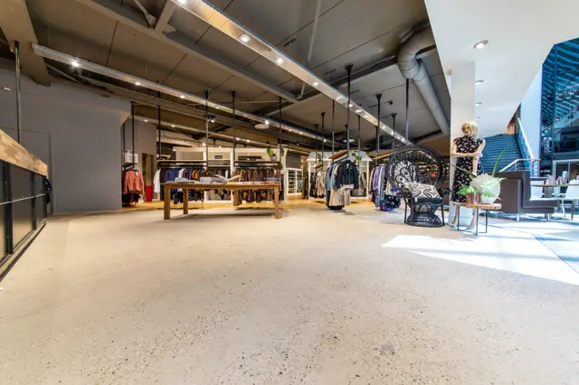 Ruime, lichte winkelruimte met een groot naadloos vloeroppervlak in warme, natuurlijke tint. De vloer creëert een uniforme, elegante basis voor de geëxposeerde kledingcollectie en houten displaytafels.