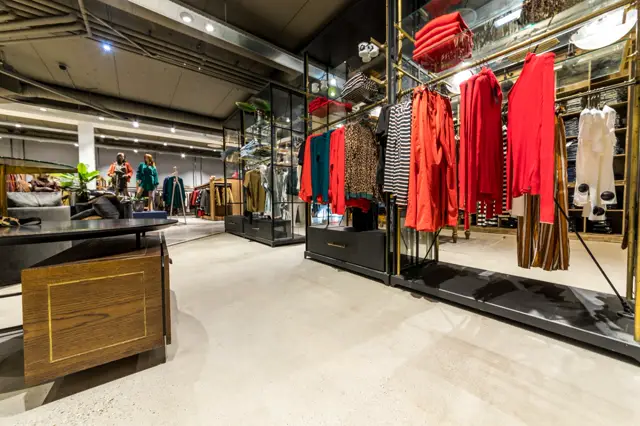 Ruime, moderne winkelruimte met heldere naadloze vloer. Gestileerde kledingdisplays met rode kledingstukken tegen gouden frames; minimalistisch opgezet interieur met professionele belichting.