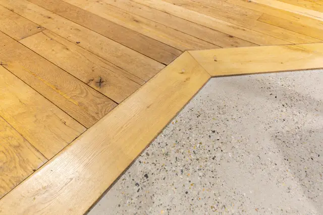 Detail van een elegante overgang tussen houten visgraat vloer en een lichte terrazzo naadloze vloer. De scherpe lijn en perfecte afwerking tonen vakmanschap en design.