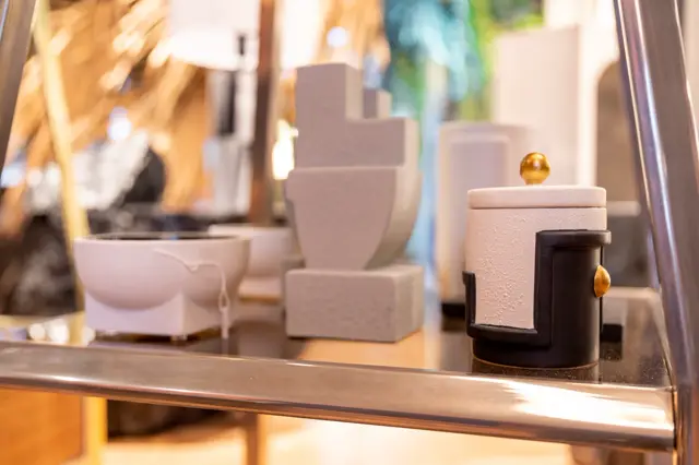 Stijlvolle display van minimalistische sculpturen op een glanzende, naadloze wand. Geometrische vormen in crème en zwart met gouden accenten, perfect uitgelicht met warm daglicht.