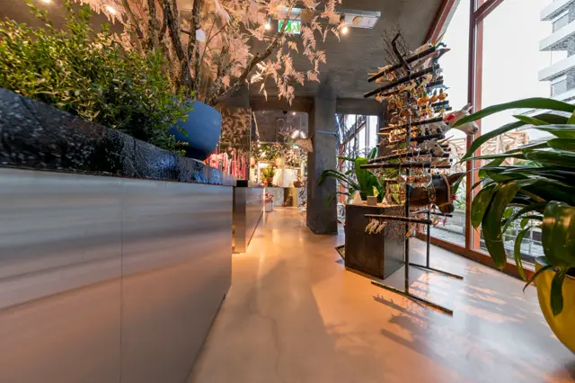 Een strakke, lichte naadloze vloer loopt door een galerijachtige ruimte met gelaagde plantenstelling en geëxposeerde kunstwerken. Warme spotverlichting accentueert de continuïteit van de vloer.