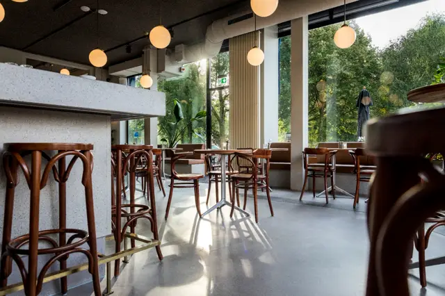Lichte, open cafézaal met naadloze vloer in warme grijstint. Grote ramen bieden uitzicht op groen, elegante hangende verlichting en houten stoelen creëren een verfijnde sfeer.