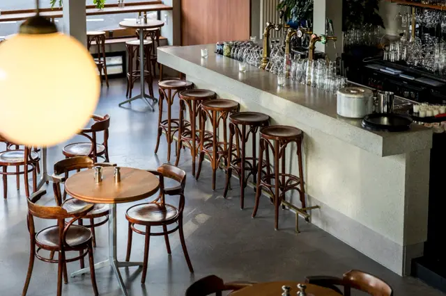 Een naadloze, licht gekleurde bartafel met scherpe lijnen domineert deze luxe café-ruimte. De vloer is donker en spiegelglad, met houten barkrukken en klassieke café-inrichting die rust en verfijning uitstralen.