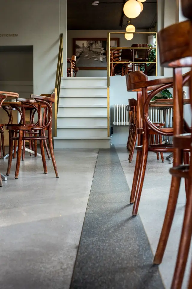 Een strakke, lichte ruimte met een naadloze vloer die twee niveaus verbindt. De vloer loopt elegant onder de trap door, omgeven door vintage houten stoelen en warme verlichting.