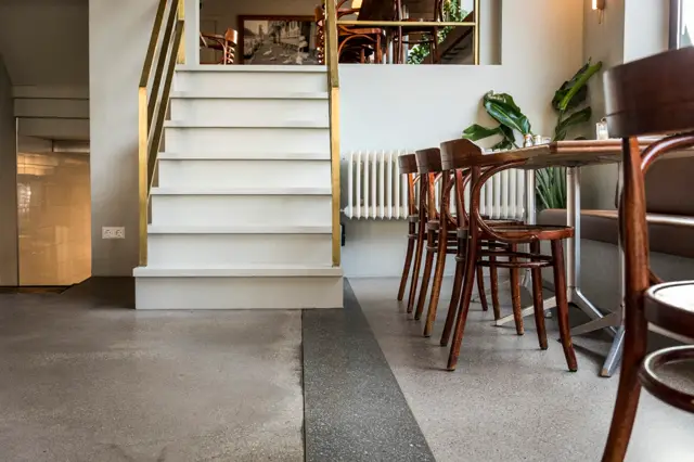 Strakke, minimalistische hal met naadloze vloer in twee grijstinten. Witte trap met messing leuning, witte muren, houten stoelen en plant. Perfecte harmonie tussen licht, ruimte en afwerking.