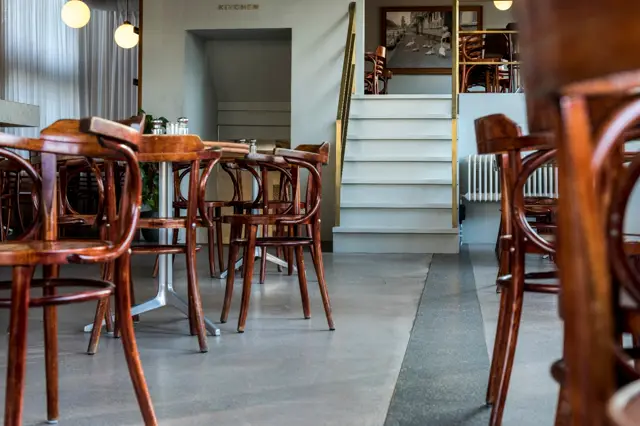 Strakke keukenruimte met lichtgrijze naadloze vloer die elegant doorloopt naar het eetgedeelte. Klassieke houten stoelen contrasteren subtiel met de moderne, schone architectuur en warme pendelverlichting.