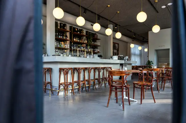 Een strak, licht café met lichtgrijze naadloze vloer die de ruimte groter maakt. Witte bar met houten stoelen, warmwitte hangende bollen en schone, rechte lijnen creëren een rustige, moderne sfeer.