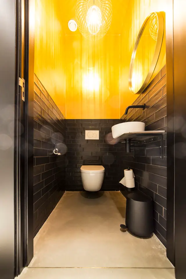 Een minimalistisch toilet met naadloze beige vloer, zwarte tegels aan de zijwanden en een opvallende gele accentwand boven met geometrische verlichting. Alle elementen zijn strak uitgelijnd en smetteloos.