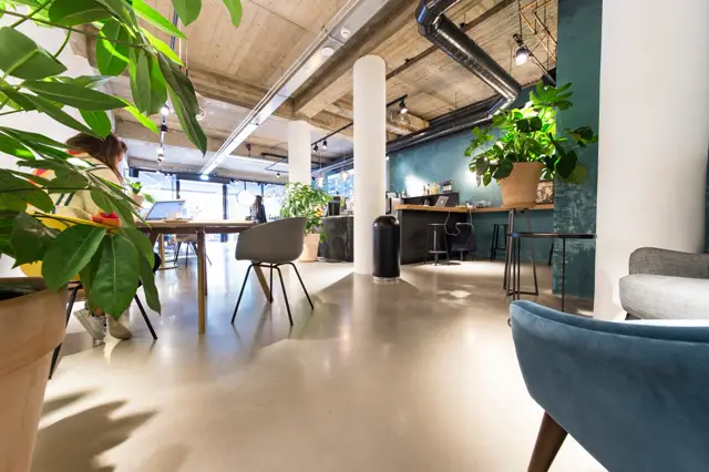 Ruim, licht openplan kantoor met strakke naadloze vloer in warm beige-grijs. Industriële plafondstructuur en groenblauwe accentwanden creëren een contrastrijke, opgeruimde werksfeer.