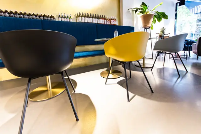 Een stijlvolle hospitality-ruimte met een heldere, naadloze vloer in warm grijs. Minimalistisch design met zwarte en gele stoelen, gouden accenten en een blauwe bar op de achtergrond, perfect verlicht door natuurlijk licht.