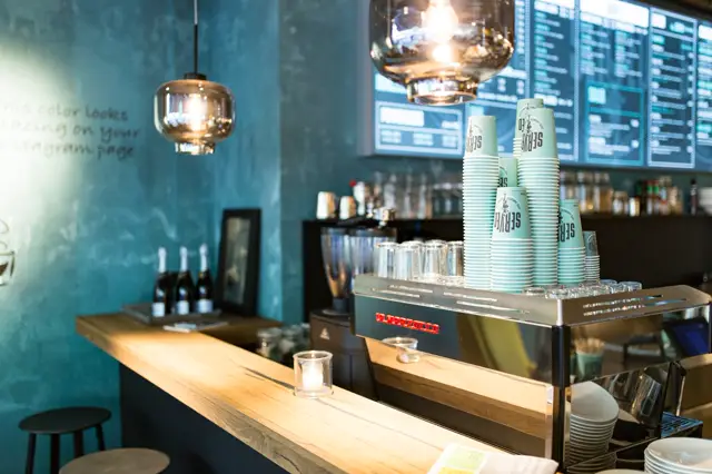 Een stijlvolle koffiecounter met een opvallende turquoise naadloze wand, glanzende hangende lampen en houten balie. Het interieur ademt rust en minimalisme uit met scherpe lijnen en doordachte sfeer.