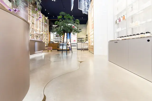 Luxe winkelruimte met strakke witte wanden en een glanzende, naadloze vloer in licht beige. Minimalistisch ingedeelde schappen en een groen plantje in het centrum creëren een airy, opgeruimd interieur.