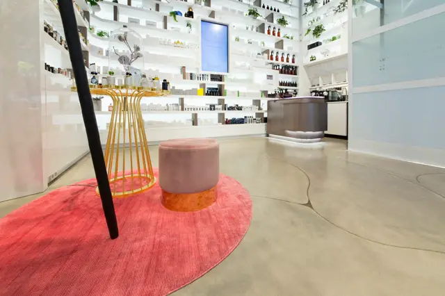 Schoon, modern retailinterieur met grote naadloze vloer in warm beige. Witte wandkasten, gouden designelement, roze accenttapijt en strak afgewerkte balie creëren een galerie-achtige uitstraling.