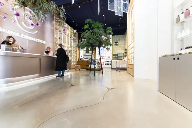 Luxe cosmeticawinkel met een volledig naadloze, lichte vloer die elegant doorloopt en visueel rust geeft. Het interieur combineert minimalistisch design met organische elementen: groene planten, heldere belichting en gestructureerde witte wandpanelen.
