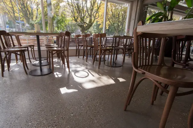Licht café-interieur met naadloze vloer in grijs-beige, natuurlijk licht van grote ramen, houten stoelen en tafels. Rustige, opgeruimde sfeer met bladgroen uitzicht.