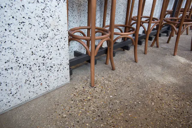 Strakke witte naadloze wand met zwarte terrazzo-accenten naast houten stoelen in een commerciële ruimte. De vloer toont een grauw, onafgewerkt oppervlak met steenslag.