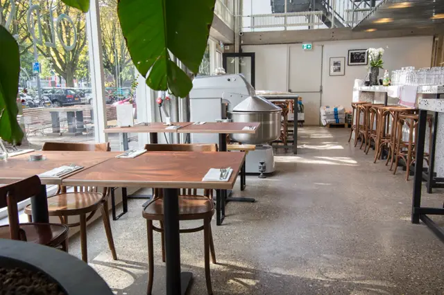 Ruim restaurant met naadloze vloer in warm grijs, doorstroomd door natuurlicht. Schone, minimalistische uitstraling met houten tafels en stalen accenten.