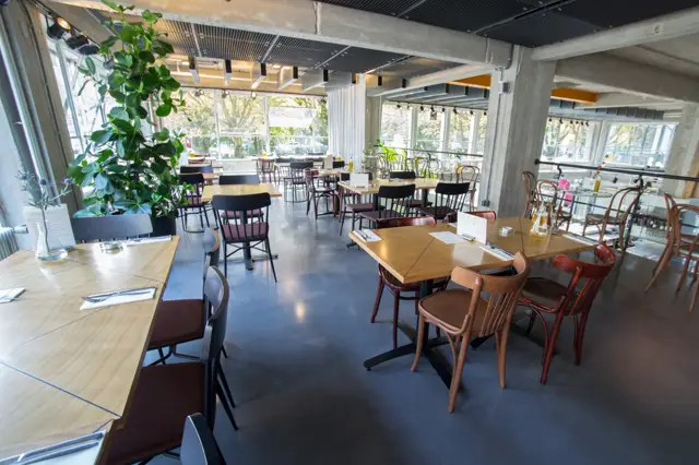 Een licht, modern restaurant met grote ramen, houten tafels en een donkere naadloze vloer die de hele ruimte visueel verbindt. Schone lijnen, groen accent en uitnodigende atmosfeer.