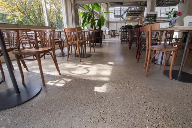 Ruime, lichte cafetariaruimte met naadloze vloer in warm grijs. Natuurlijk zonlicht valt in scherpe lijnen over het oppervlak; minimalistische houten stoelen en groene planten versterken de strakke, opgeruimde uitstraling.