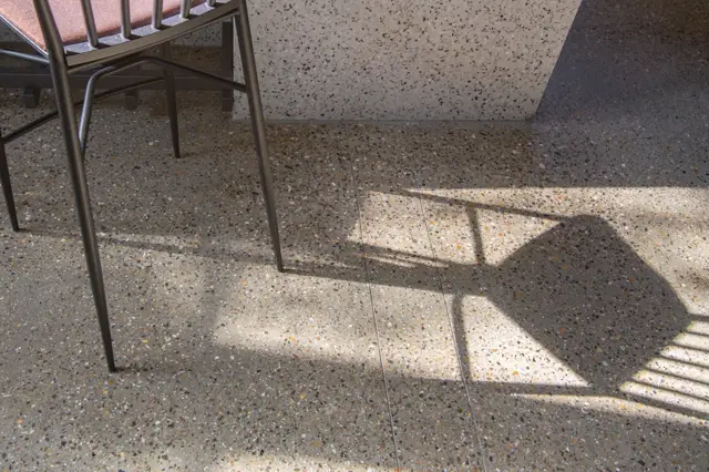 Een elegant uitgevoerde naadloze vloer in neutrale tinten met verfijnde vlekkeringen, subtiel geaccentueerd door natuurlijk zonlicht dat scherpe schaduwen van een minimalistische stoel werpt.