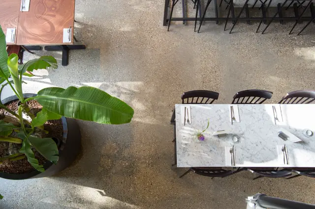 Vogelperspectiefopname van een luxe buitenterras met naadloze vloer en elegant gedekte tafels. Zonlicht creëert subtiele schaduwen; tropische planten en minimalistisch design onderstrepen de rustige sfeer.