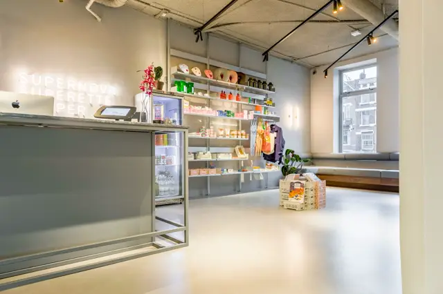 Licht, strak ingerichte beautywinkel met gladdde naadloze vloer in warm wit. Minimalistisch interieur met houten stellages, groene balie en grote ramen voor natuurlijk licht.