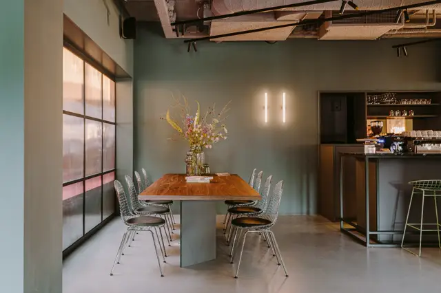 Stijlvolle kantoor- of showroomruimte met matte groene wand, natuurlijk licht van grote ramen, houten tafel met designstoelen en naadloze grijsbeige vloer. Een verfijnde sfeer met minimalistisch interieur.
