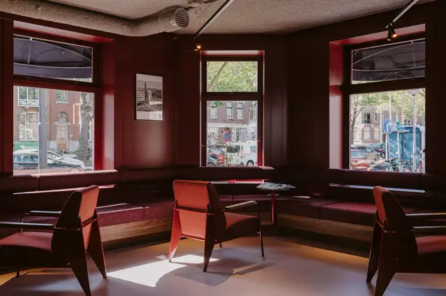 Stijlvolle bedrijfsruimte met dieprode wanden, grote ramen met natuurlicht en een naadloze vloer in warme tonen. Moderne stoelen en geometrische lijnvoering creëren een verfijnde, zakelijke sfeer.