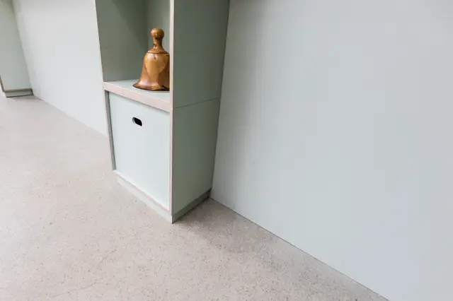 Een strak ontworpen hal met naadloze lichte vloer en witte wanden, onderbroken door een groen accent. Een houten decoratief object op de ingebouwde nis vormt een warme tegenkleur in dit serene, geordende interieur.