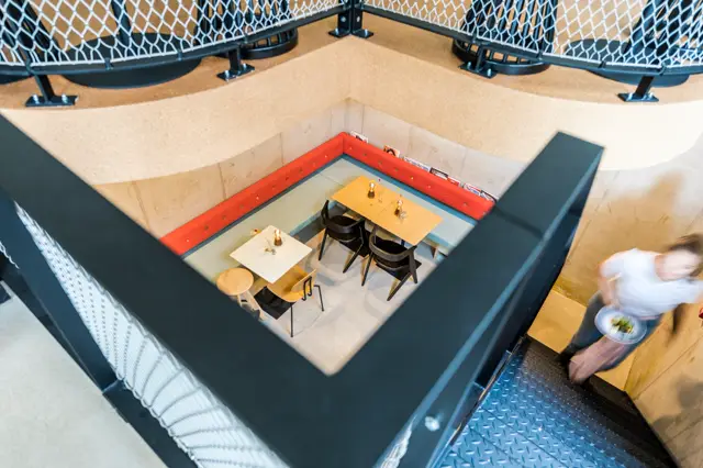 Vogelvluchtfoto van een moderne ondergrondse eetruimte met een strakke, lichte naadloze vloer. De ruimte heeft minimalistisch design met houten tafels, zwarte stoelen en kleuraccentenwanden in rood en geel.