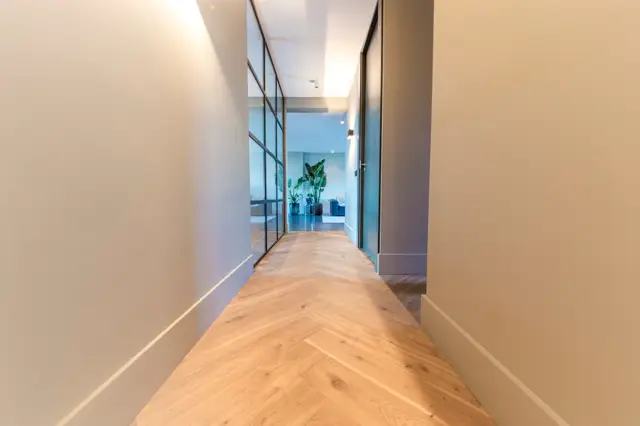 Een sierlijke, rechte gang met lichte naadloze vloer in houtlook en strakke witte wanden. Natuurlijk licht valt door glazen wand naar een aangrenzende ruimte met groen accent.
