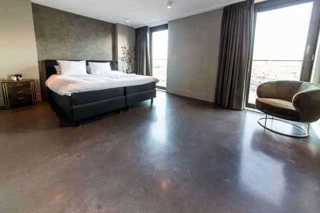 Ruime master bedroom met gladde, donkere naadloze vloer die het licht reflecteert. Strak design met neutrale wanden, minimalistisch meubilair en panoramische ramen met uitzicht.