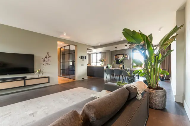 Ruime, lichte woonkamer met naadloze donkere vloer, strakke witte wanden en plafond, minimalistisch ingericht met natuurlijke planten en doordachte verlichting. Het open plan met keuken erachter straalt rust en luxe uit.